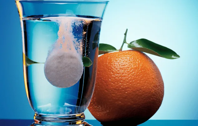 A vitamin C effervescent tablet melting in a clear glass of water next to an orange.