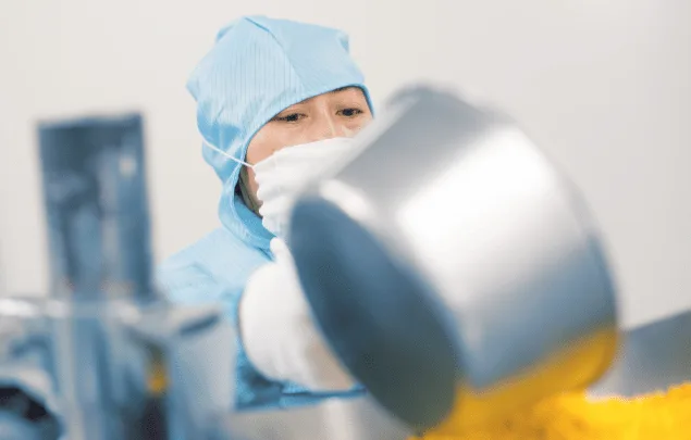 A rotary tablet press operator wearing PPE pouring powder overhead into a hopper.